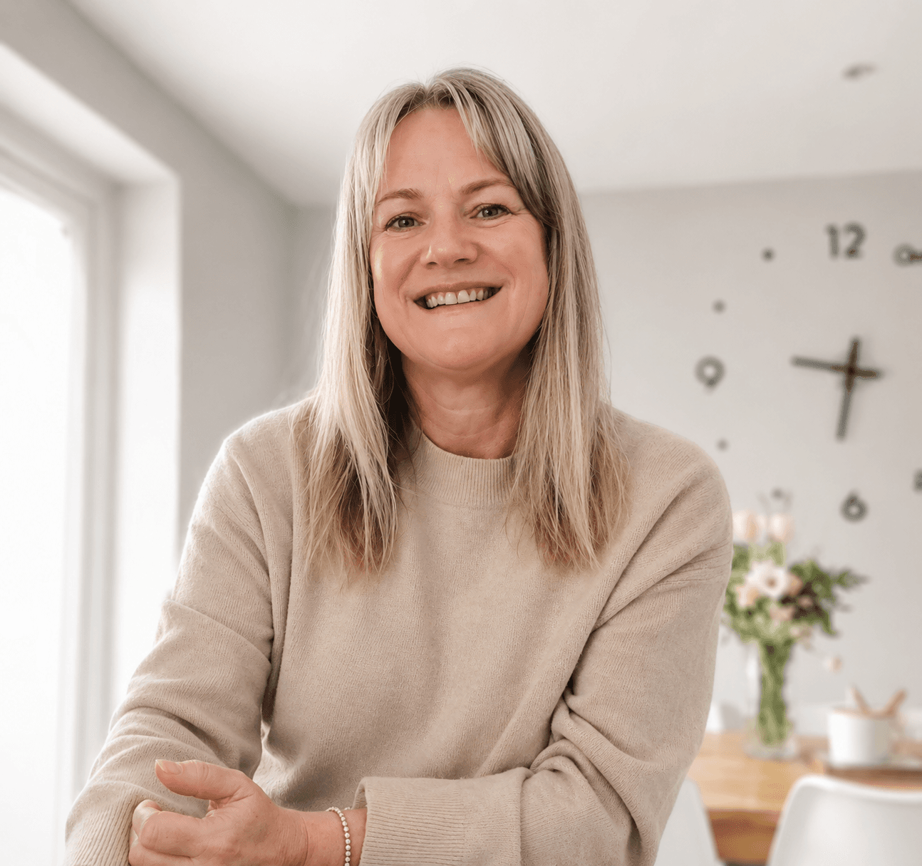 Smiling middle-aged woman with blonde hair wearing a beige sweater in a bright room.